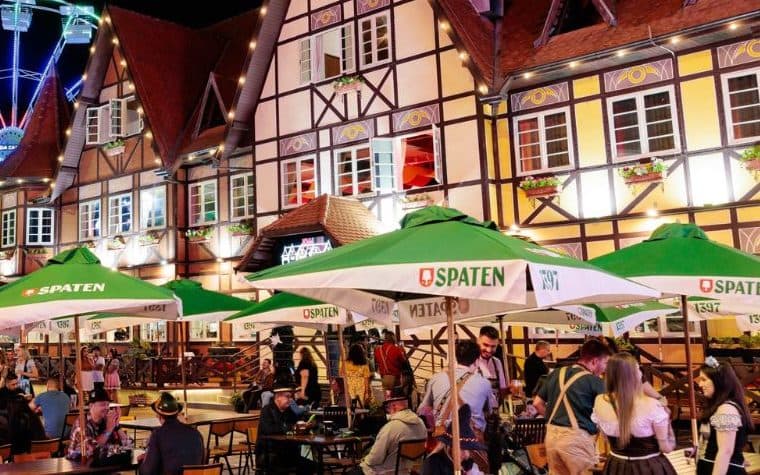 Oktoberfest de Blumenau à noite: prédios de arquitetura enxaimel iluminados no Parque Vila Germânica, com mesas ao ar livre e público em trajes típicos
