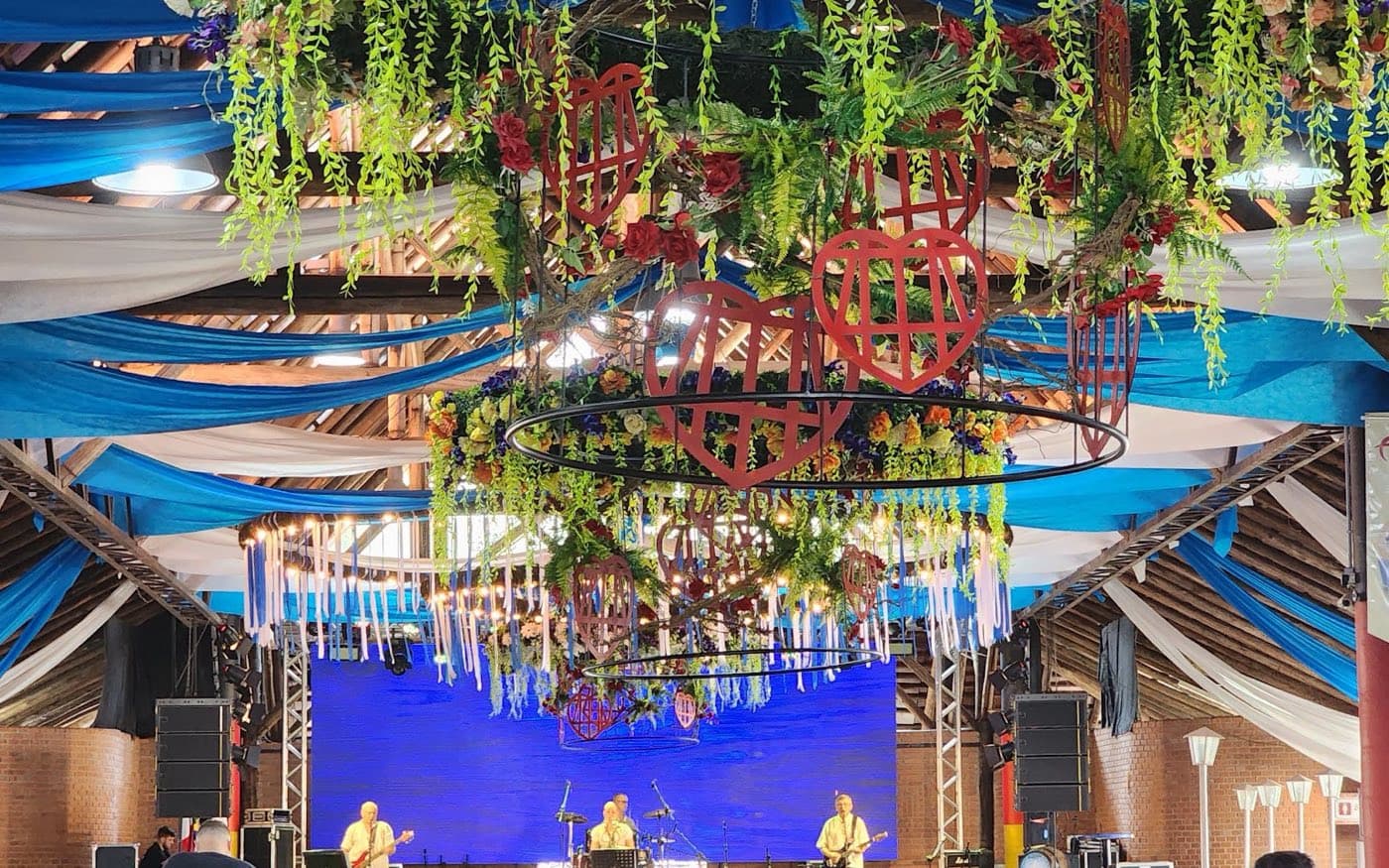 Pavilhão da Festa Pomerana decorado com fitas azuis e brancas, flores suspensas e corações vermelhos, com banda no palco ao fundo